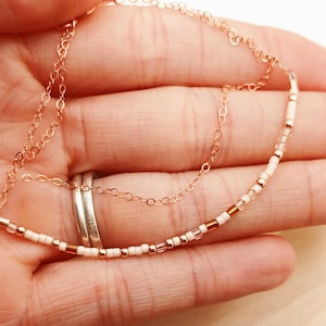 May include: Delicate rose gold necklace with a beaded strand of white and copper-colored beads. The necklace is held in a hand, with a second, simple chain visible. The text "The sister I chose" is visible in the background.