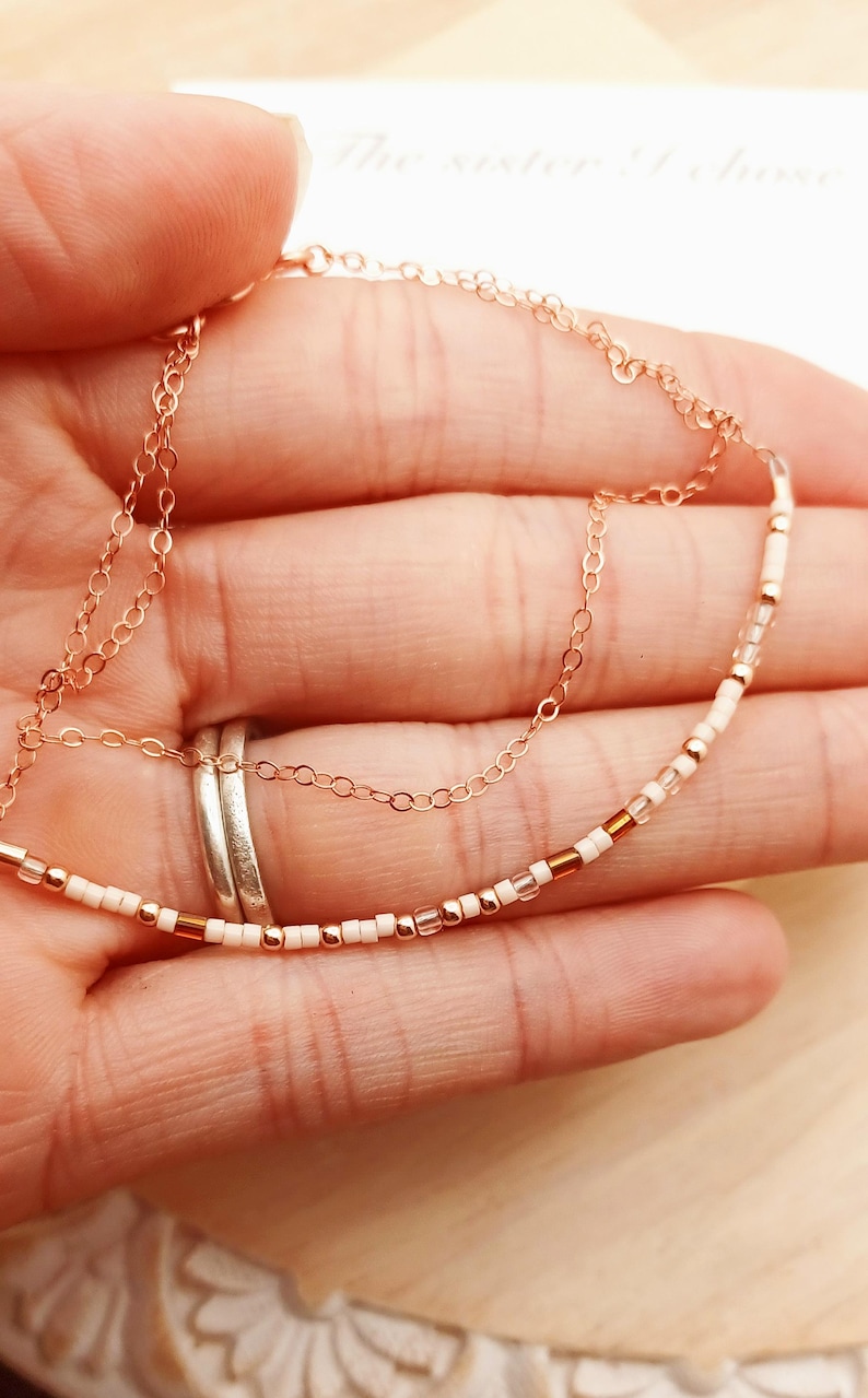 May include: Delicate rose gold necklace with a beaded strand of white and copper-colored beads. The necklace is held in a hand, with a second, simple chain visible. The text "The sister I chose" is visible in the background.