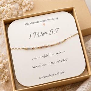 May include: A gold chain bracelet featuring a Morse code pattern made of small metallic and cream-colored beads. The bracelet is displayed on a white card printed with the text 1 Peter 5:7, Morse Code - 14k Gold Filled, and timelesselegance3.com.