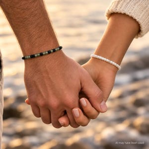 May include: Two beaded bracelets worn on wrists. One bracelet features alternating matte black and translucent olive green cylindrical beads. The second bracelet consists of a single strand of small, uniform, glossy white spherical beads.