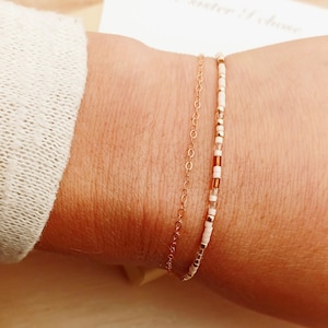 May include: Two delicate bracelets on a wrist. One is a gold chain, the other is a beaded bracelet with white, gold, and brown beads. A white card with the text "The sister I chose" is in the background.