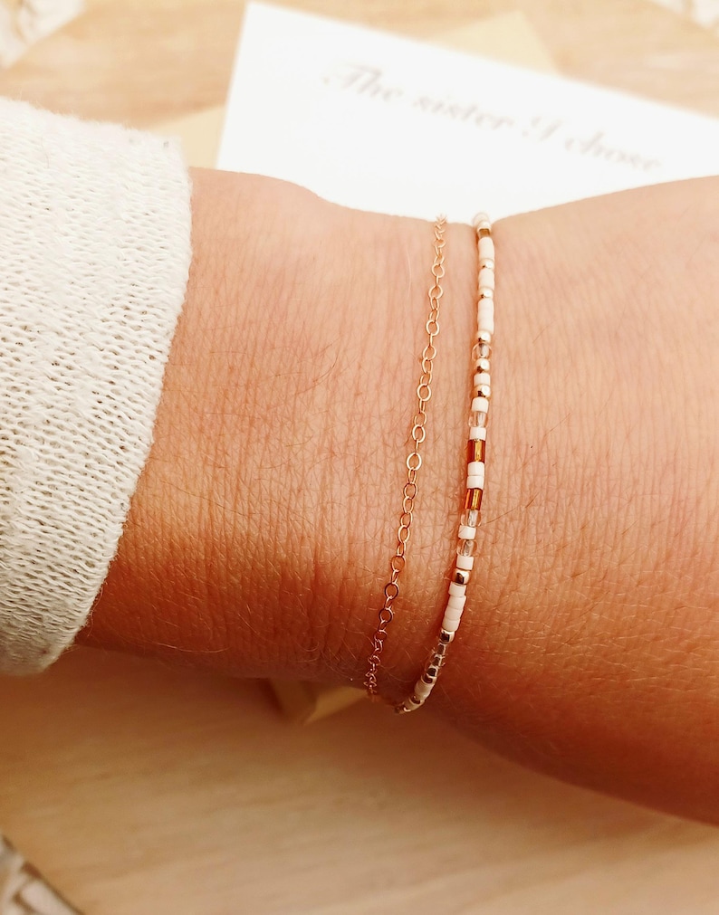 May include: Two delicate bracelets on a wrist. One is a gold chain, the other is a beaded bracelet with white, gold, and brown beads. A white card with the text "The sister I chose" is in the background.