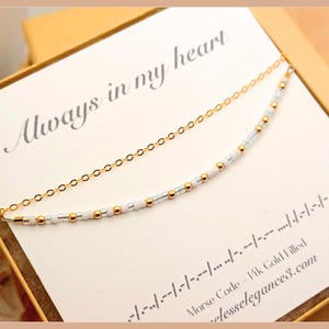 以下が含まれることがあります： モールス信号のパターンで配置された小さな白とメタリックゴールドのビーズの列が特徴の繊細なゴールドチェーンネックレス。ネックレスは、Always in my heartというテキストとモールス信号の記号が印刷された白いカードに展示されています。