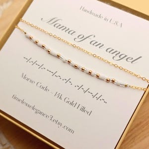 以下が含まれることがあります： 14金ゴールドフィルドのチェーンネックレスで、白とメタリックゴールドのビーズがモールス信号のパターンで並んでいます。ネックレスは、Mama of an angelという文字とモールス信号の詳細が印刷された白いカードの上に展示されています。