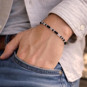 May include: Black and clear beaded bracelet. The bracelet features small, round black beads interspersed with clear, cylindrical beads. The bracelet is worn on a wrist, with a light-colored shirt and blue jeans in the background.