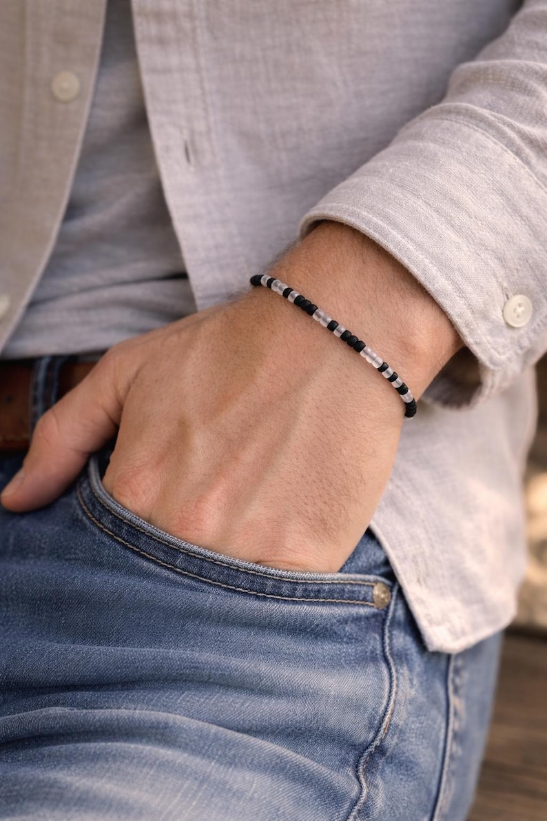 May include: Black and clear beaded bracelet. The bracelet features small, round black beads interspersed with clear, cylindrical beads. The bracelet is worn on a wrist, with a light-colored shirt and blue jeans in the background.