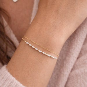 May include: A delicate gold bracelet with small white beads and gold accents is shown on a wrist. A second, similar bracelet is also visible. A heart-shaped pendant necklace is in the background. Jewelry.