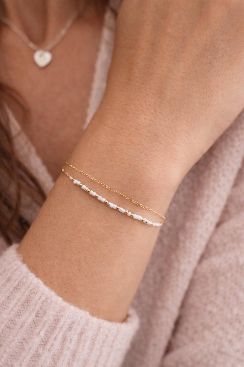 May include: A delicate gold bracelet with small white beads and gold accents is shown on a wrist. A second, similar bracelet is also visible. A heart-shaped pendant necklace is in the background. Jewelry.