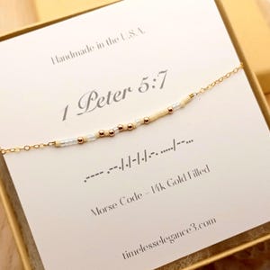 May include: A gold-filled Morse code bracelet with a delicate chain, presented on a card inside a gold box. The card reads "1 Peter 5:7" and "Morse Code - 14k Gold Filled." The website timelesselegance3.com is also visible.