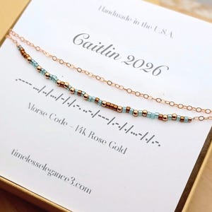 May include: A rose gold necklace with a delicate chain and a Morse code design. The necklace features small, colorful beads spelling out "Caitlin 2026". The card reads "Handmade in the U.S.A." and "timelesselegance3.com".
