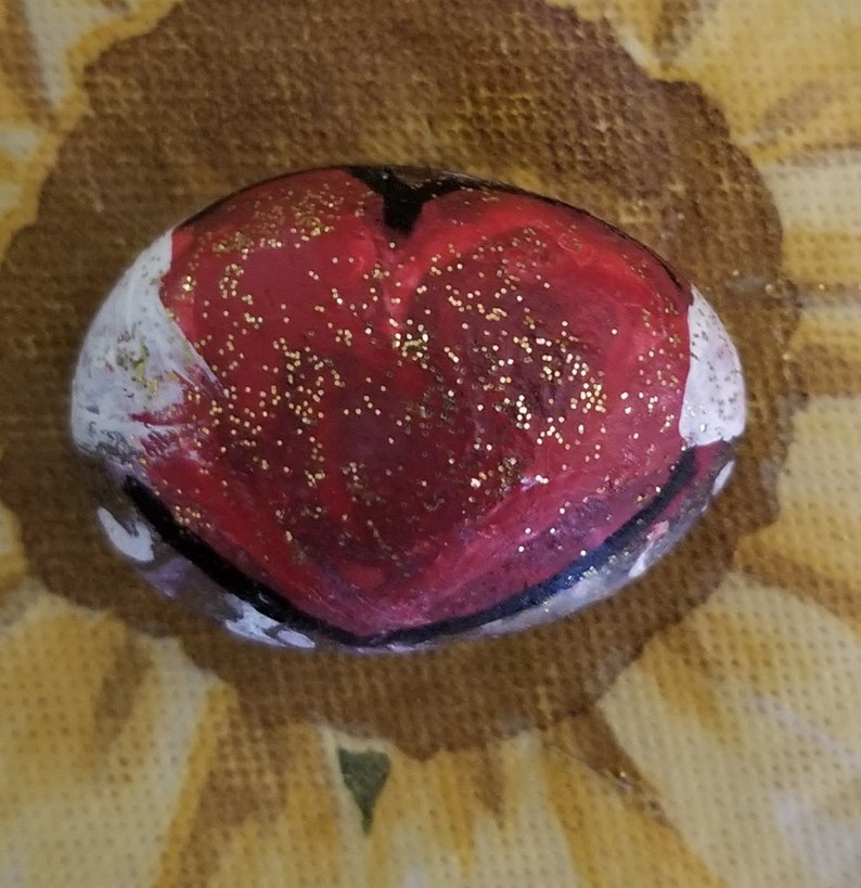 Puede incluir: Una piedra pintada con un dise&ntilde;o de coraz&oacute;n rojo y purpurina dorada. El coraz&oacute;n est&aacute; delineado en negro y la piedra tiene acentos blancos.