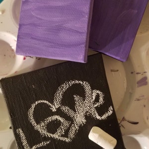 Puede incluir: Tres lienzos peque&ntilde;os, dos pintados de morado y uno negro con la palabra "Love" escrita en tiza blanca. Un trozo de tiza blanca est&aacute; sobre el lienzo negro.