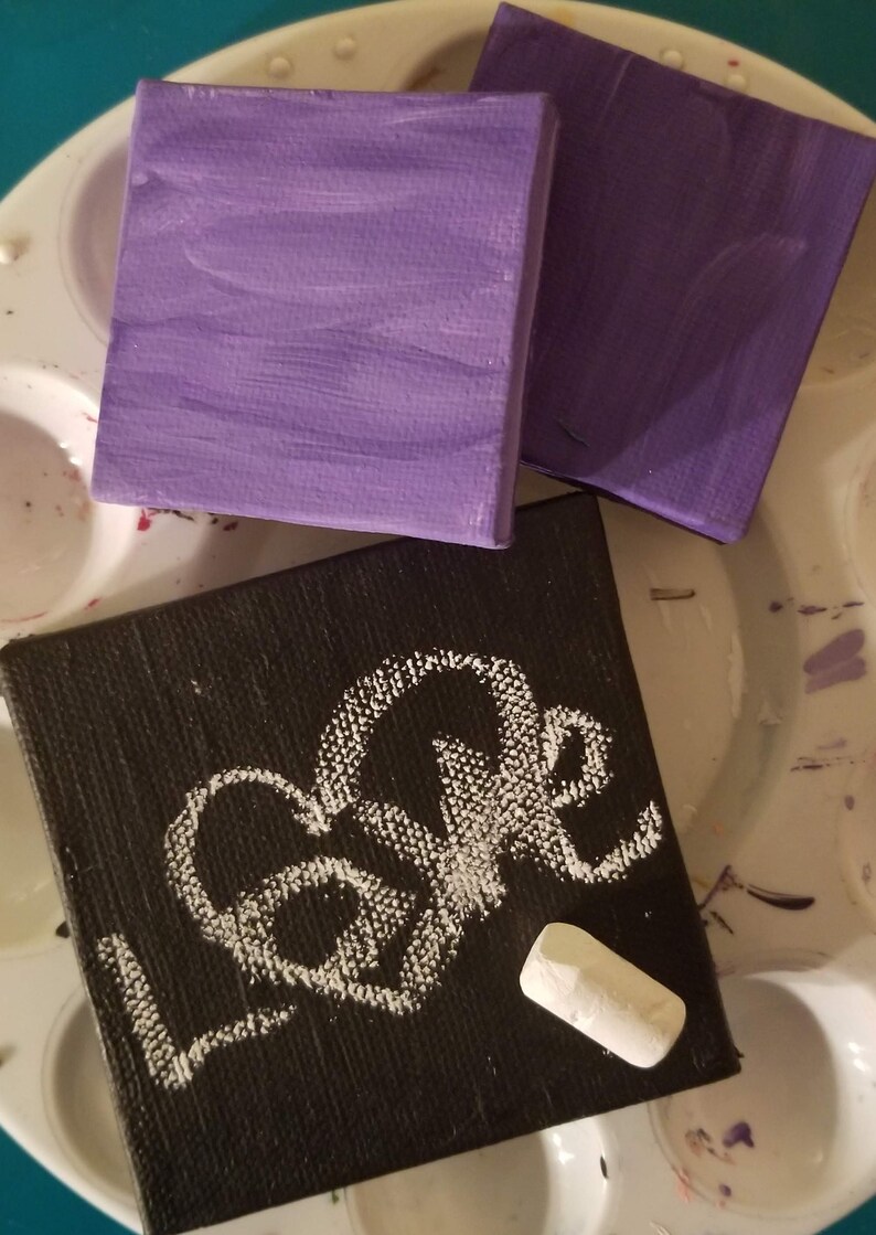 Puede incluir: Tres lienzos peque&ntilde;os, dos pintados de morado y uno negro con la palabra "Love" escrita en tiza blanca. Un trozo de tiza blanca est&aacute; sobre el lienzo negro.