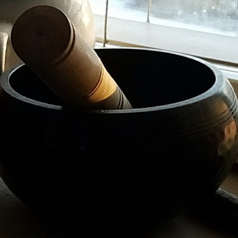 Puede incluir: Un cuenco cantor negro con un mazo de madera. El cuenco tiene una superficie lisa y redondeada y un labio ligeramente acampanado. El mazo est&aacute; hecho de madera y tiene un extremo redondeado.