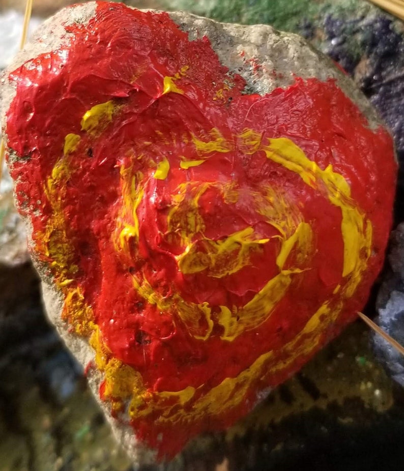 Puede incluir: Primer plano de una roca pintada con pintura roja y amarilla. La pintura se aplica en un patr&oacute;n de remolino, creando un dise&ntilde;o vibrante y abstracto.