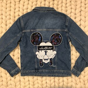 Op de afbeelding: Een blauwe denim jas met een galaxy print Mickey Mouse ontwerp op de rug. De Mickey Mouse bedekt zijn ogen met zijn handen en de tekst "whatever" staat over zijn ogen.