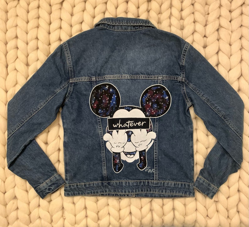 Op de afbeelding: Een blauwe denim jas met een galaxy print Mickey Mouse ontwerp op de rug. De Mickey Mouse bedekt zijn ogen met zijn handen en de tekst "whatever" staat over zijn ogen.