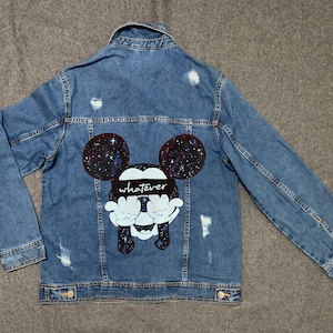 Op de afbeelding: Een blauwe denimjas met een distressed look. De achterkant van de jas heeft een zwart-witte illustratie van Mickey Mouse met een galaxy print op zijn oren en het woord "whatever" over zijn ogen.