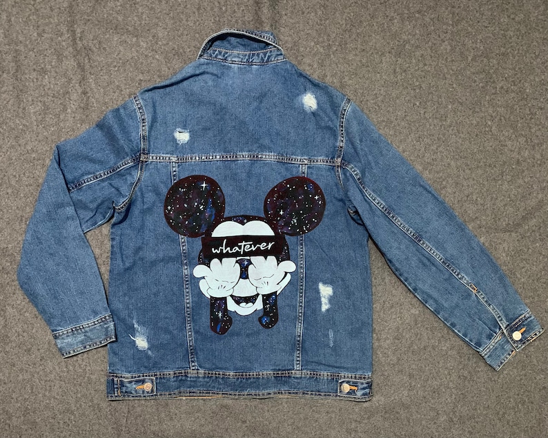 Op de afbeelding: Een blauwe denimjas met een distressed look. De achterkant van de jas heeft een zwart-witte illustratie van Mickey Mouse met een galaxy print op zijn oren en het woord "whatever" over zijn ogen.