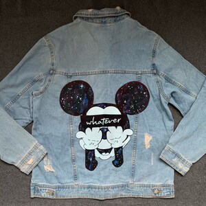 Op de afbeelding: Een lichtblauw denim jack met een handgeschilderd ontwerp van Mickey Mouse die een zwarte bandana draagt met het woord "whatever" erop gedrukt. Het ontwerp heeft een galaxy print op de oren en het hoofd van Mickey Mouse.