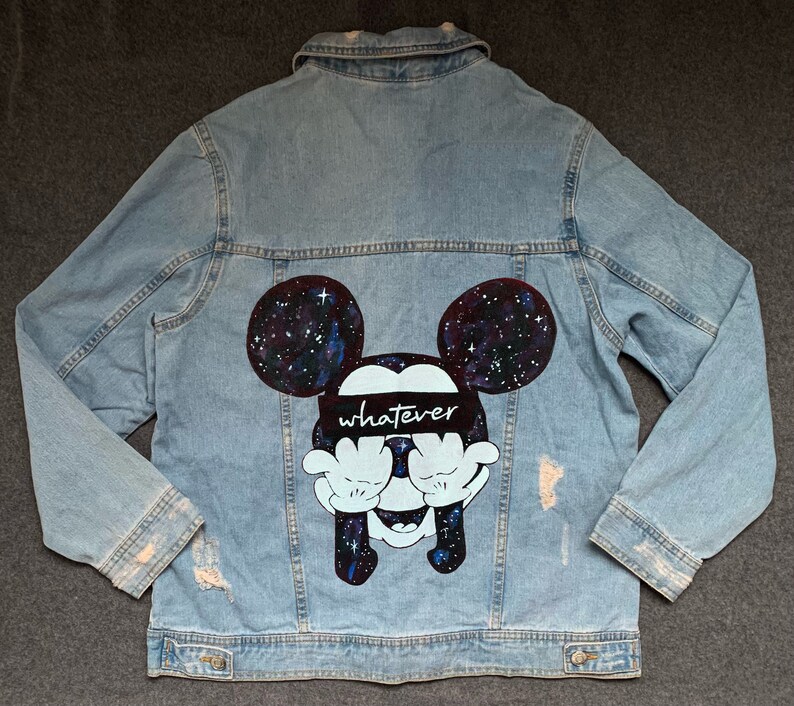 Op de afbeelding: Een lichtblauw denim jack met een handgeschilderd ontwerp van Mickey Mouse die een zwarte bandana draagt met het woord "whatever" erop gedrukt. Het ontwerp heeft een galaxy print op de oren en het hoofd van Mickey Mouse.
