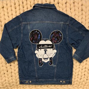 Op de afbeelding: Een blauwe denim jas met een Mickey Mouse-ontwerp met een galaxy-thema op de rug. Het ontwerp bevat het woord "whatever" over Mickey's ogen.