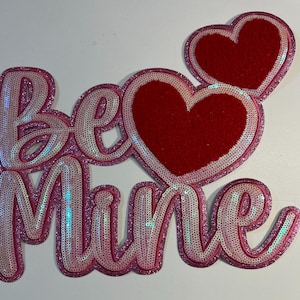 May include: Pink and red felt and sequin applique with the words "Be Mine" and two hearts.