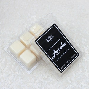 May include: A clear plastic clamshell package containing six off-white wax melt cubes. The package has a black label with the text "Light by Candle Co." and "Lavender" in white script. The label also indicates the product is soy wax and weighs 2.6 oz.