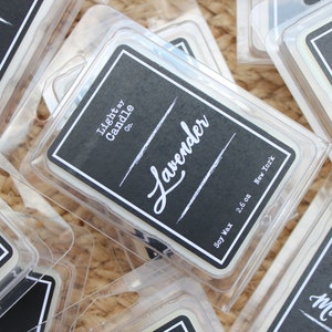Puede incluir: Primer plano de varios paquetes de cera de soja. Los paquetes est&aacute;n en recipientes de pl&aacute;stico transparente con etiquetas negras. Las etiquetas tienen texto blanco que dice "Lavender", "Light my Candle Co." y "2.6 oz New York".