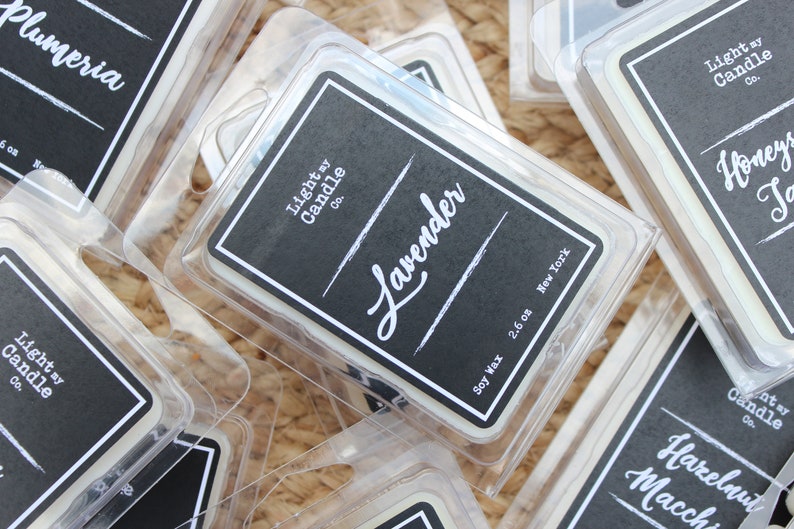 Puede incluir: Primer plano de varios paquetes de cera de soja. Los paquetes est&aacute;n en recipientes de pl&aacute;stico transparente con etiquetas negras. Las etiquetas tienen texto blanco que dice "Lavender", "Light my Candle Co." y "2.6 oz New York".