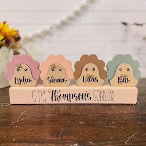 May include: Four wooden turkey figurines with painted faces and names: Lydia, Shannon, Lucas, and Ben. The turkeys are sitting on a wooden block with the text "G-Ma Thompson's Gobblers".