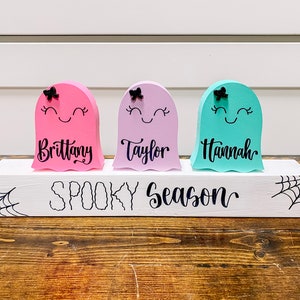 Pu&ograve; includere: Tre figurine di fantasmi in legno con volti dipinti e fiocchi, in piedi su un blocco di legno bianco con il testo "Spooky Season". I fantasmi sono dipinti di rosa, viola e turchese e hanno i nomi "Brittany", "Taylor" e "Hannah" scritti su di loro.