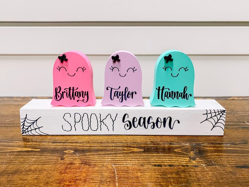 Pu&ograve; includere: Tre figurine di fantasmi in legno con volti dipinti e fiocchi, in piedi su un blocco di legno bianco con il testo "Spooky Season". I fantasmi sono dipinti di rosa, viola e turchese e hanno i nomi "Brittany", "Taylor" e "Hannah" scritti su di loro.