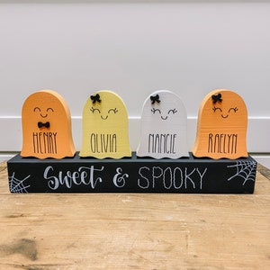 Pu&ograve; includere: Quattro figurine di fantasmi in legno con volti dipinti e fiocchi, ciascuna con un nome scritto sotto: Henry, Olivia, Nancy e Raelyn. I fantasmi sono seduti su una base in legno nero con le parole "Sweet & Spooky" scritte in bianco.