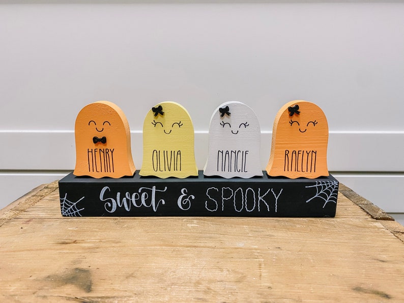 Pu&ograve; includere: Quattro figurine di fantasmi in legno con volti dipinti e fiocchi, ciascuna con un nome scritto sotto: Henry, Olivia, Nancy e Raelyn. I fantasmi sono seduti su una base in legno nero con le parole "Sweet & Spooky" scritte in bianco.