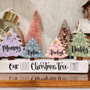 May include: A white wooden sign with the text "Our Little Christmas Tree Farm" painted in black. Four small wooden Christmas trees are attached to the sign, each painted in a different color and with a name written on it: "Mommy", "Ayden", "Averleigh", and "Daddy".