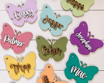 May include: Ten wooden butterfly ornaments with different names written on them. The butterflies are painted in various colors, including pink, purple, green, yellow, blue, and orange. The names on the butterflies are: Emery, Jagger, Palmer, Cohen, Tripp, Mae, Maddox, Brack, and Josephine.