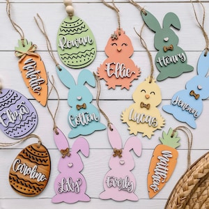 Może przedstawiać: R&oacute;żne drewniane ozdoby wielkanocne w pastelowych kolorach, w tym kr&oacute;liki, kurczaki, marchewki i jajka. Każda ozdoba ma imię w kontrastowym kolorze i jest przewleczona sznurkiem i drewnianymi koralikami. Ozdoby znajdują się na białej drewnianej powierzchni.