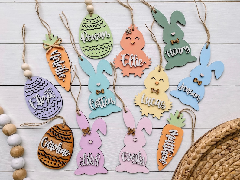 Może przedstawiać: R&oacute;żne drewniane ozdoby wielkanocne w pastelowych kolorach, w tym kr&oacute;liki, kurczaki, marchewki i jajka. Każda ozdoba ma imię w kontrastowym kolorze i jest przewleczona sznurkiem i drewnianymi koralikami. Ozdoby znajdują się na białej drewnianej powierzchni.