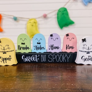Pu&ograve; includere: Un set di sei figurine di fantasmi in legno con volti dipinti e nomi. I fantasmi sono di vari colori, tra cui giallo, verde, blu, viola, rosa e bianco. I fantasmi sono disposti su un cartello di lavagna nera che recita "Sweet But Spooky".