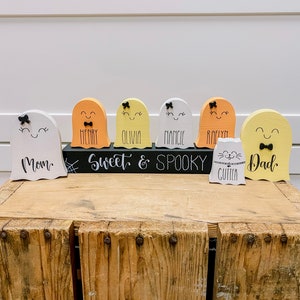 Pu&ograve; includere: Un set di sette figurine di fantasmi in legno con volti dipinti e fiocchi. I fantasmi sono dipinti in varie tonalit&agrave; di giallo, arancione e bianco. I fantasmi sono disposti in fila su un cartello nero che recita "Sweet & Spooky". I fantasmi sono etichettati con nomi tra cui "Mom", "Henry", "Olivia", "Mancie", "Roelyn", "Dad" e "Cutter".