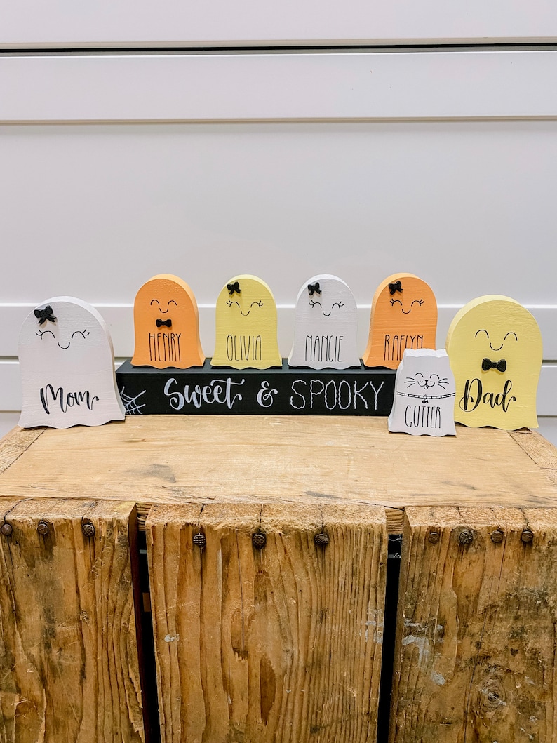 Pu&ograve; includere: Un set di sette figurine di fantasmi in legno con volti dipinti e fiocchi. I fantasmi sono dipinti in varie tonalit&agrave; di giallo, arancione e bianco. I fantasmi sono disposti in fila su un cartello nero che recita "Sweet & Spooky". I fantasmi sono etichettati con nomi tra cui "Mom", "Henry", "Olivia", "Mancie", "Roelyn", "Dad" e "Cutter".