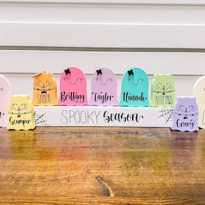 Pu&ograve; includere: Un cartello di legno bianco con il testo "Spooky Season" in lettere nere. Il cartello &egrave; decorato con ragnatele nere e otto figurine di fantasmi colorati con nomi scritti su di esse. I fantasmi sono di colore rosa, arancione, giallo, verde, blu, viola e bianco.