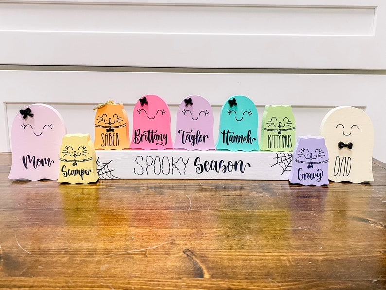 Pu&ograve; includere: Un cartello di legno bianco con il testo "Spooky Season" in lettere nere. Il cartello &egrave; decorato con ragnatele nere e otto figurine di fantasmi colorati con nomi scritti su di esse. I fantasmi sono di colore rosa, arancione, giallo, verde, blu, viola e bianco.