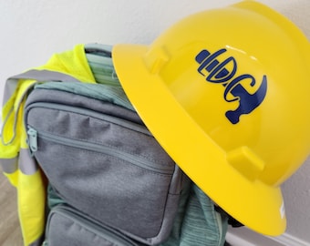 May include: A yellow construction helmet with a blue logo rests atop a gray backpack. A high-visibility vest and work gloves are also visible. The backpack has multiple pockets and a teal-colored front. The items suggest a construction or work-related theme.