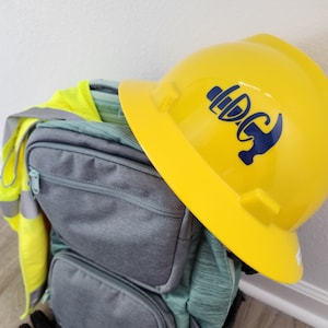 May include: A yellow construction helmet with a blue logo rests atop a gray backpack. A high-visibility vest and work gloves are also visible. The backpack has multiple pockets and a teal-colored front. The items suggest a construction or work-related theme.