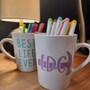 May include: Two white ceramic mugs with colorful pens inside. One mug has "BEST LIFE EVER" in teal lettering. The other mug has a purple graphic. The mugs are on a wooden surface.