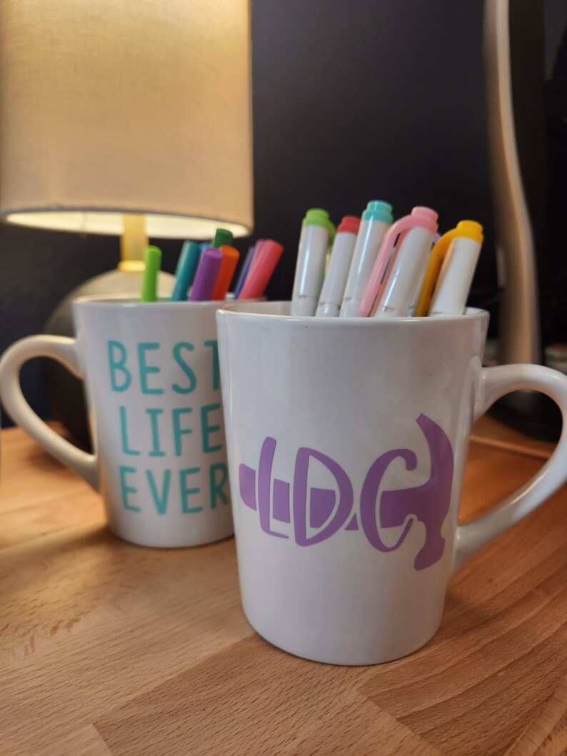 May include: Two white ceramic mugs with colorful pens inside. One mug has "BEST LIFE EVER" in teal lettering. The other mug has a purple graphic. The mugs are on a wooden surface.