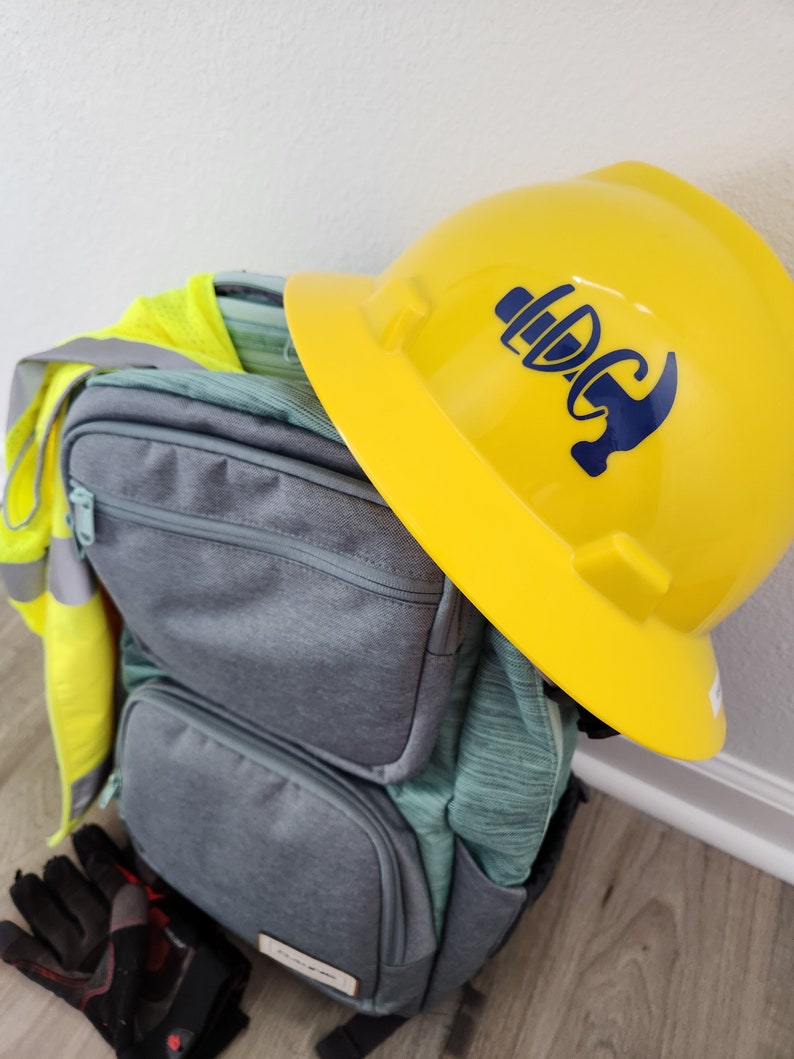 May include: A yellow construction helmet with a blue logo rests atop a gray backpack. A high-visibility vest and work gloves are also visible. The backpack has multiple pockets and a teal-colored front. The items suggest a construction or work-related theme.