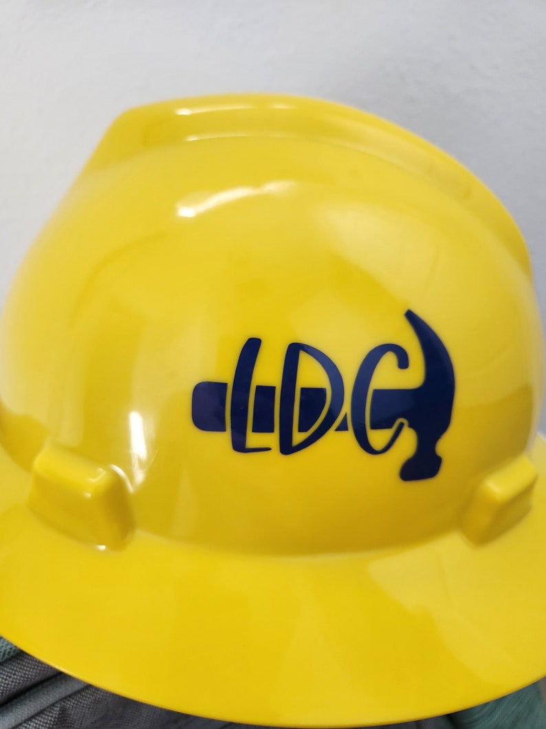 May include: A bright yellow hard hat with a navy blue logo that reads "LDC" with a hammer graphic. The hard hat is made of a glossy material and has a standard construction design.